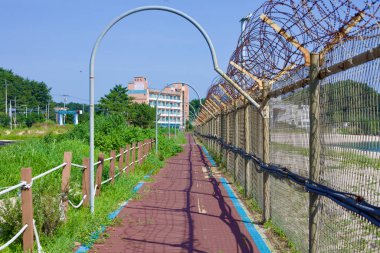 Goseong County, South Korea - July 30, 2019: A beachside bike path adorned with blue lines and arching supports, separated from the beach by old barbed wire fencing, lies near Gyoam Beach and Port, with urban buildings rising in the background. clipart