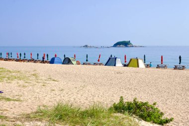 Goseong County, Güney Kore - 30 Temmuz 2019: Piknik masaları ve kapalı şemsiyeler Bongpo Sahili 'nin sahilinde, arkasında çadır kurarak, Doğu Denizi' ndeki Jukdo Adası 'nın arka planına karşı bir rahatlama ve rahatlama sahnesi kuruyorlar..
