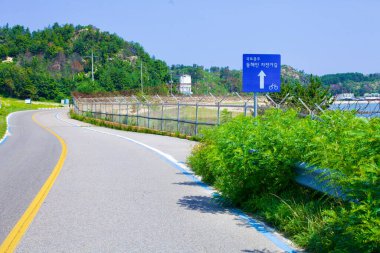 Goseong County, South Korea - July 30, 2019: Alongside a road near the north end of Gonghyeonjin Beach, a bike path sign and a distinct blue line indicate the route as part of the Gangwon bike path, with barbed wire-topped fencing separating the path clipart