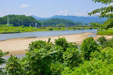 Goseong County, South Korea - July 30, 2019: Gazing upriver, the Bukcheon Railroad Bridge emerges in the distance, spanning a stream with prominent sandbanks, all set against a backdrop of verdant green mountains. clipart