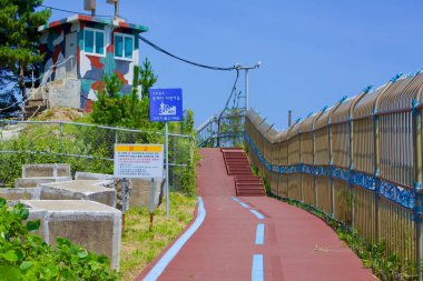 Goseong County, South Korea - July 30, 2019: A bike path, delineated by blue lines, guides to a staircase and ramps beside a camouflaged lookout tower, with a wrought iron fence on one side, set under a clear blue sky near Geojin Village Beach. clipart