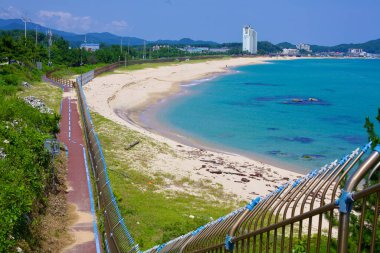 Goseong County, South Korea - July 30, 2019: From a high-angle perspective, Geojin Village Beach unfolds with its pristine sandy shores and blue waters. A bike path, bordered by a metal fence separating it from the beach, offers a scenic route, with  clipart