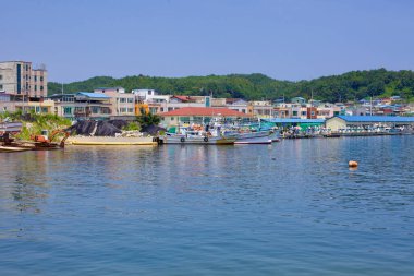 Goseong County, Güney Kore - 30 Temmuz 2019: Sergi sırasında Geojin Limanı 'nda liman boyunca demirlemiş tekneler, berrak mavi bir gökyüzünün altında, berrak mavi suları yansıtan yeşil tepelerin güzel bir zemin üzerine kurulmuştur.