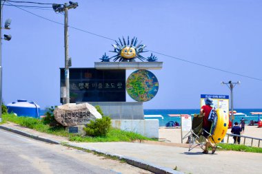 Goseong County, Güney Kore - 31 Temmuz 2019 Hwajinpo Sahili 'nin kuzey ucunda bir ziyaretçi deniz kenarında bir gün için hazırlanıyor, üzerinde bir güneş heykeli olan granit bir levhayı geçiyor, Chodo Limanı ve plaja doğru gidiyor..