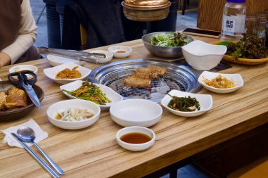 Ulsan, Güney Kore - 24 Şubat 2024: Kimçi, fasulye lahanası, sirke ve soya soslu soğan ve peçetede duran kaşık ve çubuklarla çevrili kömür ızgarasında et ızgarası