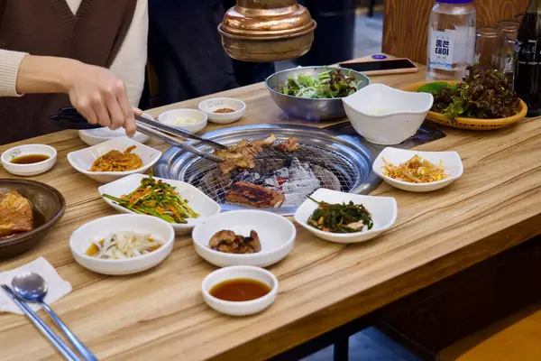 Ulsan, Güney Kore - 24 Şubat 2024: Bir samgyeopsal restoranında etrafını saran bir dizi banchan tabağıyla çevrili dumanla mangal ızgarasında sigara içilen eti ters çevirmek için maşa kullanan bir kişinin geniş bir görüntüsü.