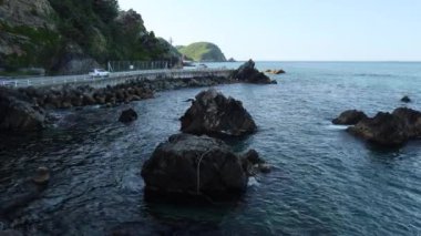 Gangneung Şehri, Güney Kore - 18 Mayıs 2024: Dalgalar Doğu Denizi 'nin sularındaki kubbeli kayalara çarpar ve Heonhwa Yolu boyunca ilerler. Arka planda, güneş tarafından öpülen yeşil sahil tepesine karşı arabalar yol boyunca gider..