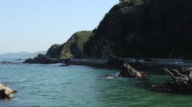 Gangneung Şehri, Güney Kore - 18 Mayıs 2024: Heonhwa Yolu 'nun durağan bir görüntüsü Doğu Denizi' nden yola çıkan bir kaya parçası. Yeşil, ağaçlarla kaplı kayalık yamaçlar dolambaçlı yoldan yükseliyor..