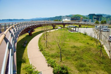 Samcheok City, Güney Kore - 18 Mayıs 2024: Isabu Plaza 'daki yüksek yürüyüş yolu ve yeşil alan manzarası. Kavisli yaya köprüsü park alanına yeni dikilmiş ağaçlar ve çevre şehir ve Doğu Denizi manzarası ile bakıyor..