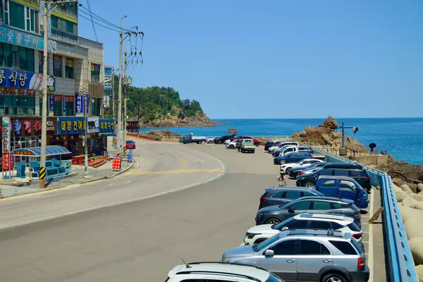 Stock image Samcheok City, South Korea - May 18th, 2024: A view towards Lion Rock from Isabu Plaza, showcasing the coastal road lined with parked cars and local shops. The rocky coastline and clear blue sea create a picturesque scene.