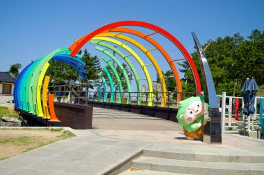 Donghae Şehri, Güney Kore - 18 Mayıs 2024: Chuam Sahili 'ndeki canlı Gökkuşağı Köprüsü, renkli kemerler ve neşeli heykeller, güneşli bir günde ziyaretçileri deniz kıyısında ağırlıyor.