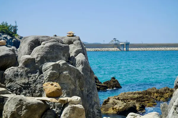 stock image Donghae City, South Korea - May 18th, 2024: View of the rocky shoreline and stone formations near Chuam Candlestick Rock in Donghae. A concrete breakwater is visible in the background, providing a contrast between natural and man-made structures. The