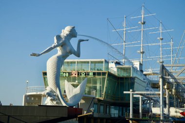 Gangneung, South Korea - May 18th, 2024: A striking mermaid statue with a water stream coming out of her mouth stands prominently on Jeongdongjin Port's breakwater, with the tall ship restaurant Fish Soup in the background. clipart