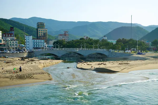stock image Gangneung, South Korea - May 18th, 2024: A picturesque view of a stream cutting through Jeongdongjin Beach, flowing under a charming bridge with the town and mountains providing a scenic backdrop.