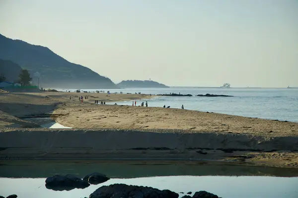 stock image Gangneung, South Korea - May 18th, 2024: Visitors stroll along the sandy shores of Jeongdongjin Beach as the sun sets, casting a serene glow over the coastline and the distant mountains.