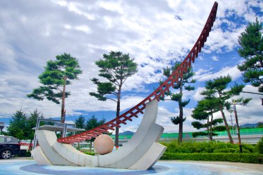 Samcheok, Güney Kore - 29 Temmuz 2024: Samcheok Deniz Demiryolu Bisiklet İstasyonu 'nun dışındaki modern bir heykel, bir çam ağacı zeminine ve parçalı bulutlu gökyüzüne karşı pürüzsüz, eğimli tasarım unsurları barındırıyor..