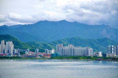 Sokcho, South Korea - July 28th, 2024: A broad view of Sokcho's urban skyline set against the rugged Taebaek Mountains, with the calm waters of Cheongcho Lake stretching across the foreground on a partly cloudy day. clipart