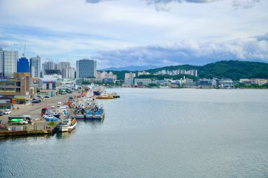 Sokcho, Güney Kore - 28 Temmuz 2024: Sokcho Limanı 'na demirlemiş balıkçı tekneleri, bulutlu bir gökyüzü altında şehrin dinamik bir görüntüsünü oluşturan modern binalar ve tepelerin arka planıyla rıhtım boyunca dizilmiştir..