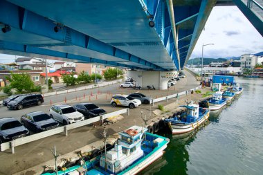 Sokcho, South Korea - July 28th, 2024: A view from beneath Geumgang Grand Bridge, showing fishing boats docked along the water and a parking area with vehicles near Abai Village in Sokcho. clipart