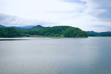 Goseong County, Güney Kore - 28 Temmuz 2024: Hwajinpo Gölü 'nün Hwajinpo Köprüsü' nden sakin suları ve Goseong County 'de bulutlu bir gökyüzünün altında yemyeşil tepeleri gösteren huzurlu bir manzara.