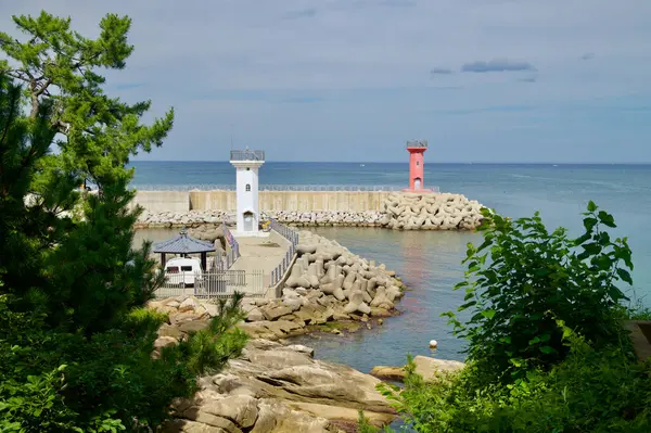 stock image Goseong County, South Korea - July 28th, 2024: The red and white lighthouses of Gyoam Port stand guard at the entrance of the breakwater, framed by lush greenery and overlooking the calm waters of the East Sea.