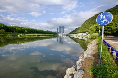 Ulsan, Güney Kore - 9 Temmuz 2019: Sakin Taehwa Nehri 'nin yanında şehrin ufuk çizgisi ve yemyeşil nehir boyunca yemyeşil manzaralı bir bisiklet ve yaya tabelası duruyor..
