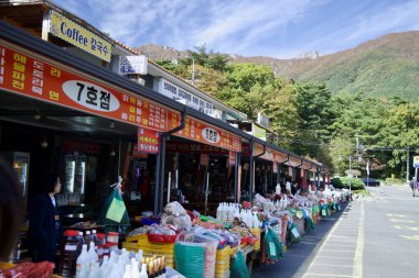 Ulsan, South Korea - October 21st, 2023: Vendors at the entrance of Seoknamsa Temple offer a variety of local goods and produce under colorful signage, with the mountainous backdrop adding to the scenic charm. clipart