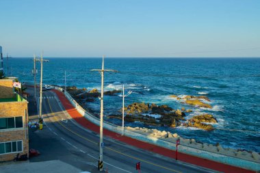 Gangneung City, South Korea - November 3rd, 2024: A serene view from Jumunjin Lighthouse overlooking a coastal road winding alongside the rocky shore of the East Sea, framed by clear blue waters. clipart