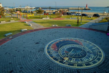 Goseong County, Güney Kore - 3 Kasım 2024: Bongpo Village Sea Wind Park 'taki plaza zasında büyük bir dekoratif saat tasarımının havadan görünüşü, arka planda Bongpo Liman feneri ve kıyı şeridi.
