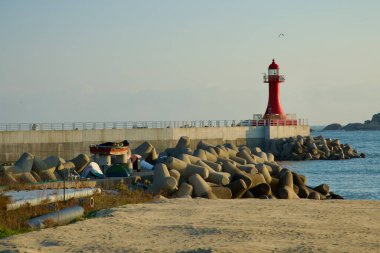 Goseong County, South Korea - November 3rd, 2024: The vibrant red lighthouse at Bongpo Port stands near a collection of tetrapods along the shore, serving as coastal protection while enhancing the maritime scenery. clipart