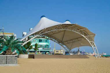 Sokcho, Güney Kore - 3 Kasım 2024: Beyaz tepe örtülü modern bir performans sahnesi, yakınlardaki palmiye ağaçları ve renkli binalar tarafından çerçevelenmiş Sokcho Sahili 'nin kumlu arazisinde yer alıyor..