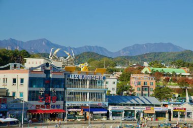 Sokcho, South Korea - November 3, 2024: Seafood restaurants line Daepo Port, featuring vibrant signage and a large crab sculpture on the rooftop, with the backdrop of scenic mountains. clipart