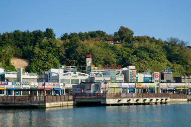 Sokcho, South Korea - November 3, 2024: A view of the waterfront at Daepo Port, featuring vibrant local shops and restaurants, with a green hillside rising in the background. clipart