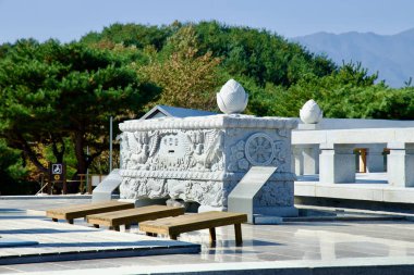 Yangyang County, South Korea - November 3rd, 2024: A detailed stone donation box adorned with intricate carvings and lotus motifs stands prominently in front of the Seawater Avalokitesvara statue at Naksansa Temple. clipart