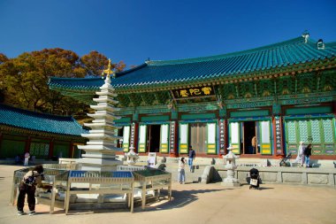 Yangyang County, South Korea - November 3rd, 2024: A serene scene of the stone pagoda standing in front of the colorful Botajeon Hall at Naksansa Temple, surrounded by autumn foliage. clipart