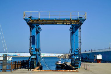 Yangyang İlçesi, Güney Kore - 3 Kasım 2024: Susan Limanı 'nda büyük mavi bir tekne asansörü, altında küçük bir gemi, canlı bir gökyüzüne ve renkli dalgalı su duvarlarına karşı kurulmuş..