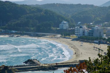 Yangyang, Güney Kore - 3 Kasım 2024: Kumlu kıyıları ve yumuşak dalgalarıyla, yemyeşil tepeler ve civarındaki modern binalarla çevrili, huzurlu bir sahil atmosferi sunan Ingu Sahili manzarası.