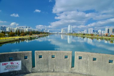 Ulsan, Güney Kore - 14 Kasım 2024: Taehwa Nehri 'nin panoramik manzarası, Hakseong Köprüsü' ndeki gözcü platformundan parçalı bulutlu sonbahar gökyüzü altında çekilmiş Ulsan 'ın modern siluetini yansıtıyor.