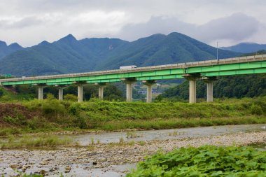Goesan County, Güney Kore - 10 Eylül 2020: Yeonpung yakınlarındaki sığ bir nehir boyunca uzanan yeşil bir ekspres köprü boyunca bir kamyon seyahat ederken, Chungcheongbuk-do 'nun dağlık manzarası manzaralı bir zemin oluşturur..