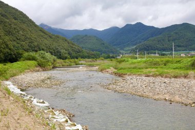 Goesan County, Güney Kore - 10 Eylül 2020: Arka planda yemyeşil dağlar olan bir vadi boyunca uzanan sığ bir nehir Kore kırsalının doğal güzelliğini gözler önüne seriyor..