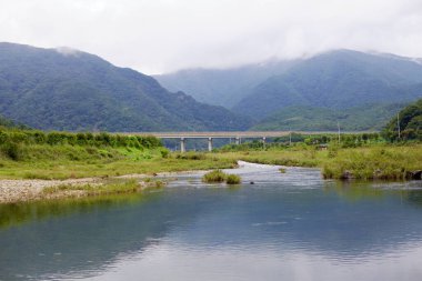 Goesan İlçesi, Güney Kore - 9 Eylül 2020: Ssang Stream Chilseong Kasabası 'ndan akar, arka planda sis kaplı dağların çerçevelediği vadi boyunca uzanan bir köprü olarak bulutlu gökyüzünü yansıtır..