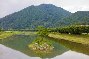 Goesan İlçesi, Güney Kore - 10 Eylül 2020: Seonghwang deresinin durgun sularında küçük, ağaçlarla kaplı bir ada, çevresindeki dağlar ve ağaçlarla çevrili kıyılar sakin yüzeye yansıyor..