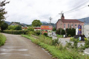 Goesan County, Güney Kore - 9 Eylül 2020: Chilseong Town 'da sessiz bir yol kenarı; kırmızı tuğlalı bir kilise, yerel işletmeler ve nemli bir patika boyunca yemyeşil bir yol, uzak mesafelerde dağların görülebileceği bir yer..