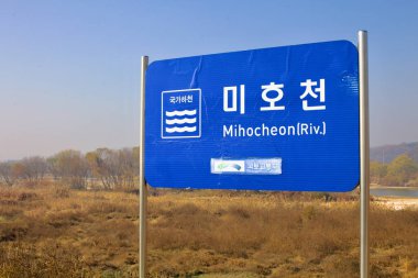Cheongju, Güney Kore - 11 Kasım 2020: Miho Stream 'in (Mihocheon) nehir kenarına yakın bir yerde olduğunu gösteren mavi bir işaret, sonbahar yaprakları ve açık bir gökyüzünün altında yuvarlanan tepeler.