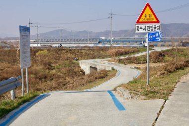 Cheongju City, South Korea - November 11, 2020: A winding section of the Ocheon Bicycle Path featuring warning signs cautioning cyclists, a narrow concrete bridge, and dry vegetation with industrial buildings in the background. clipart