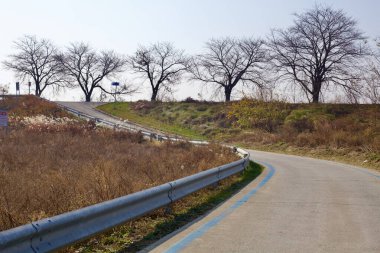 Cheongju Şehri, Güney Kore - 11 Kasım 2020: Ocheon Bisiklet Yolu 'nun bariyerler, kurumuş çimenler ve çıplak ağaçlarla kaplı dolambaçlı bir bölümü, ovalardan daha yüksek bir sete geçişi işaret ediyor..