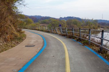 Daejeon, Güney Kore - 12 Kasım 2020: Geum Nehri boyunca metal korkuluklar ve sonbahar yapraklarıyla çevrili mavi şeritli bir bisiklet yolu manzaralı ve huzurlu bir yolculuk sunuyor..
