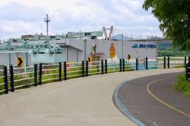 Sejong City, Güney Kore - 27 Mayıs 2021: Geum Nehri 'ndeki önemli bir su yönetim yapısı olan Sejong Weir yakınlarındaki bir kontrol tesisi etrafında bir bisiklet yolu kıvrımlı. Weir akışı düzenler ve yerel sulamayı destekler.