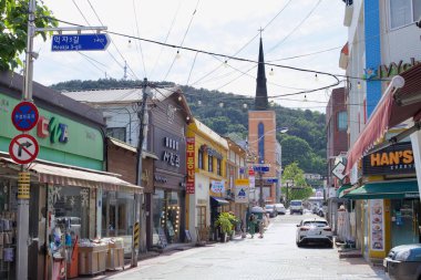 A street view of Meokja 3-gil, lined with small shops, cafs, and restaurants, leading towards a church with a tall spire against the backdrop of green hills. clipart