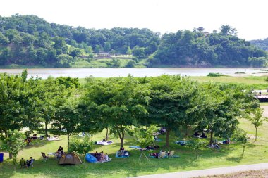 Gongju City, South Korea - May 27th, 2021: People relax in a riverside park with shaded picnic areas, overlooking the historic Gong Mountain Fortress, a UNESCO-listed Baekje-era site across the Geum River. clipart